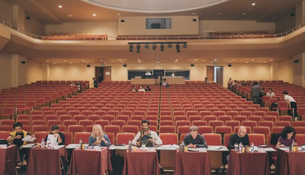 a panel of judges sitting at the front of the stage of a drama school and observing auditions 