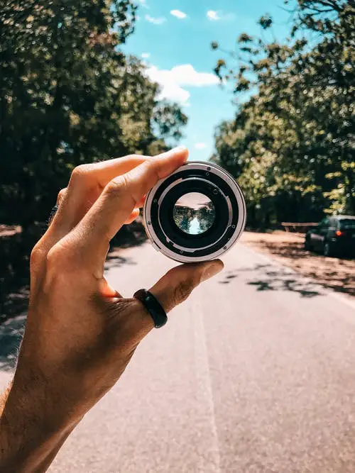 Person holding a camera length in the middle of the street under blue sky surrounded by trees
